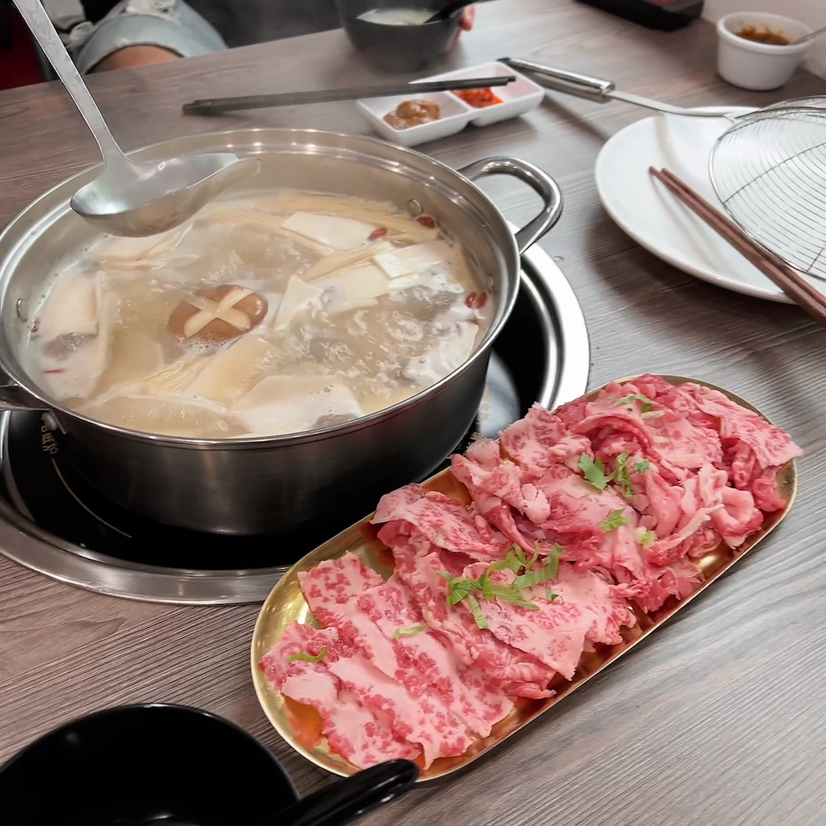 A pot of beef bone soup with mushrooms beside a gold plate of marbled sliced beef