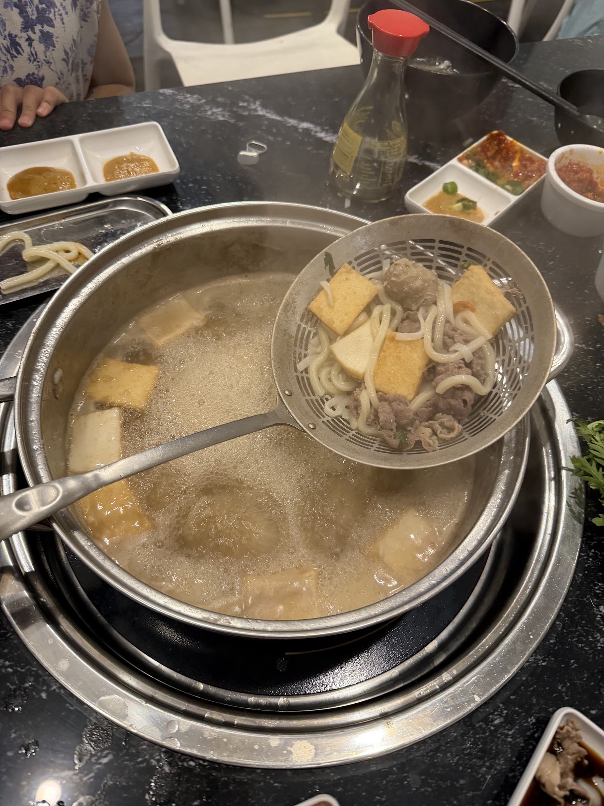 A pot of beef bone soup bubbling at the table with tofu puffs and noodles in a wire scoop