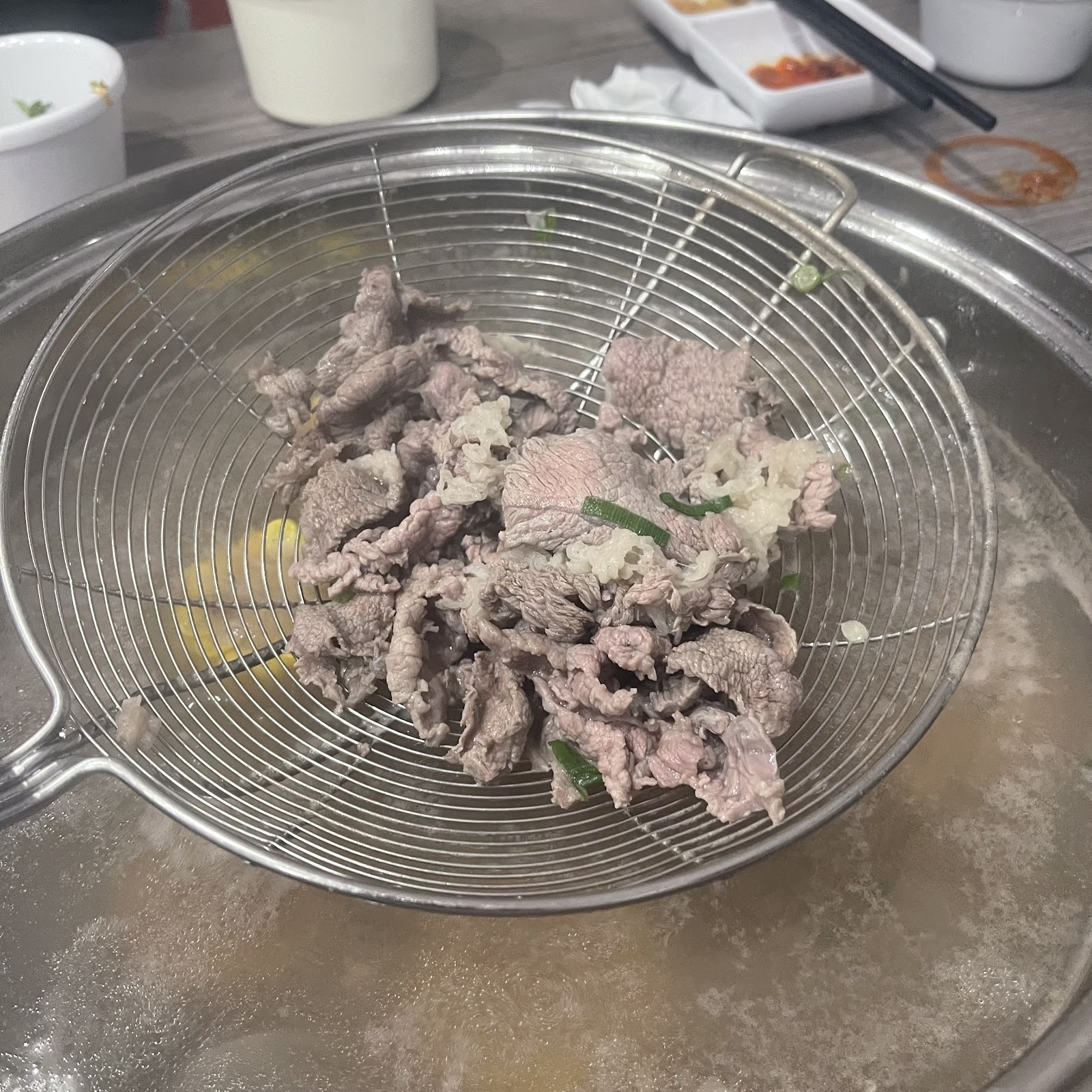 A wire scoop of freshly cooked sliced beef held above the steaming pot
